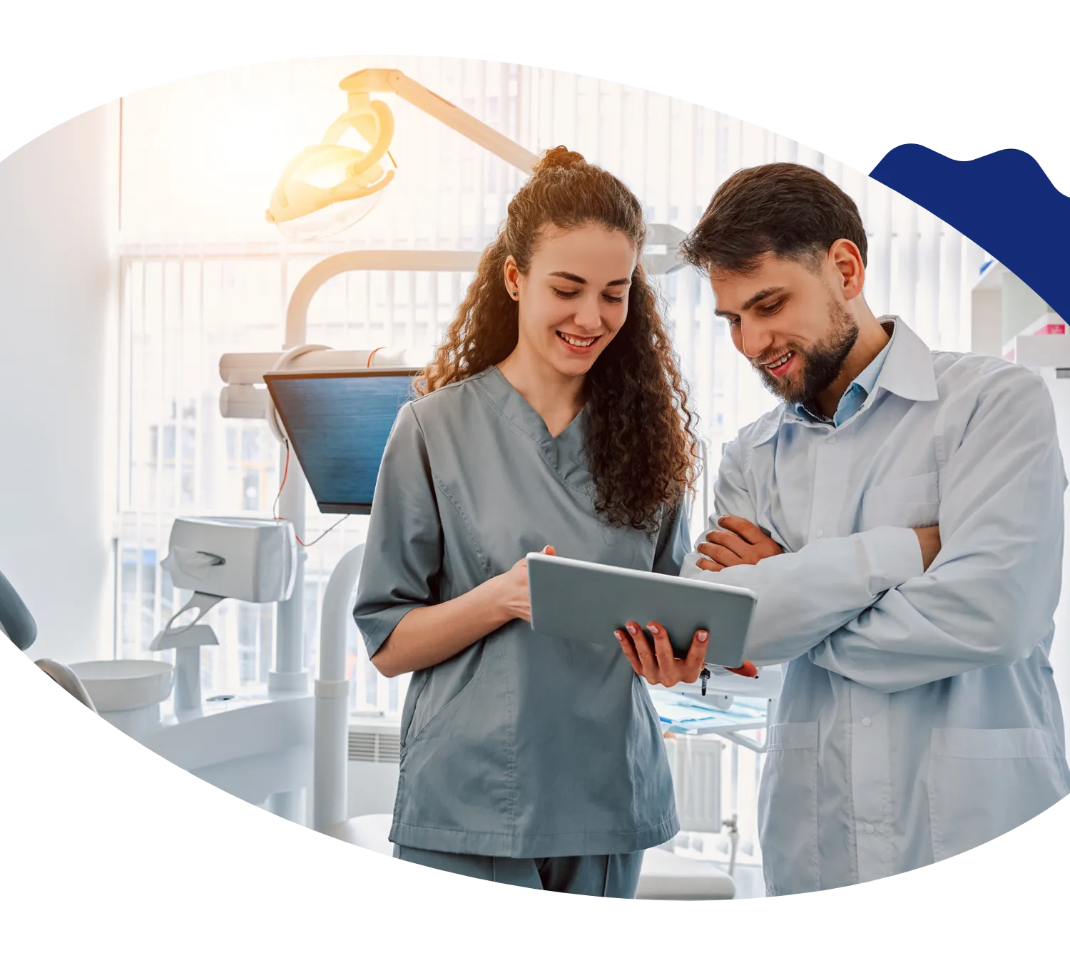 A dental professional and a hygienist review information on a tablet while standing in a modern dental office, framed within an oval-shaped design with a decoartive blue accent.